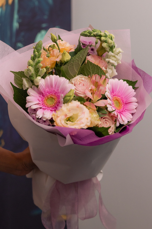 Bouquet of pastel seasonal flowers wrapped in pink paper held by a person.